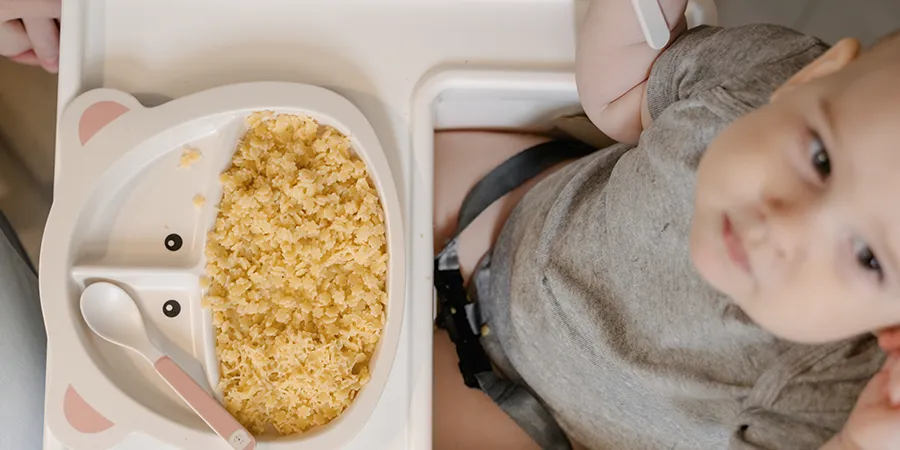 Slika odozgo bebe u hranilici. Ispred njega je zanimljivi dečiji tanjir sa hranom.