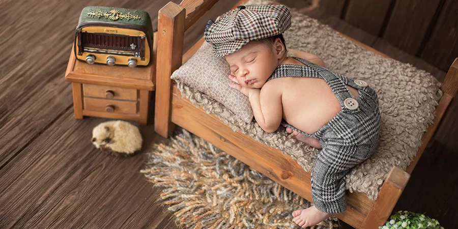 Kadar bebe odevene u karirane panralone sa tregerima i kariranom kapom na glavi. Beba spava na stomaku, na malom drvenom krevetu.
