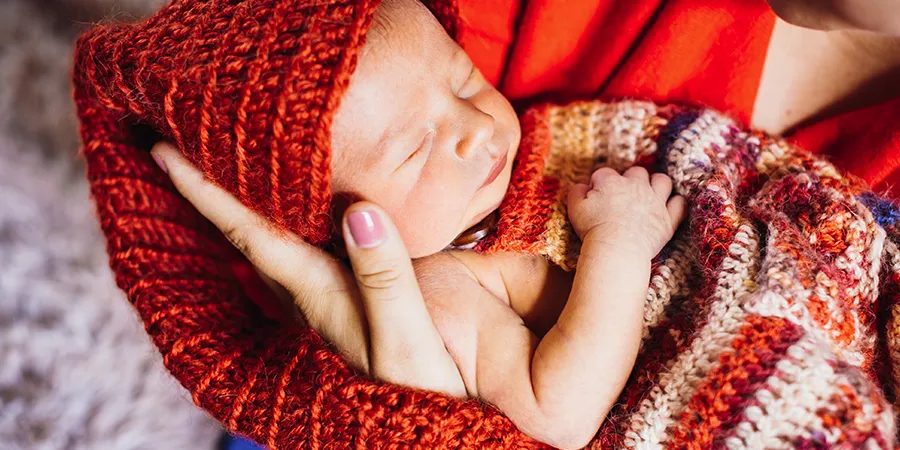 Krupni kadar novorođenčeta u maminom naručju. beba je uvijena u ručno pleteno crveno ćebe.