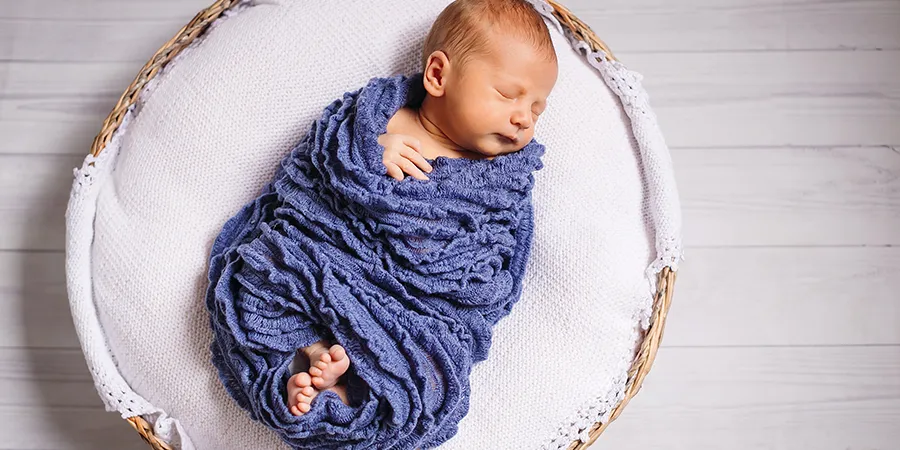 Slika odozgo bebe dečaka. uvijenog u plavo ćebe. Beba je u pletenoj korpi na belom dušeku.
