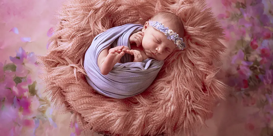 Slika odozgo bebe devojčice uvijene u ljubičasto ćebe, sa svetnom trakom na glavi.