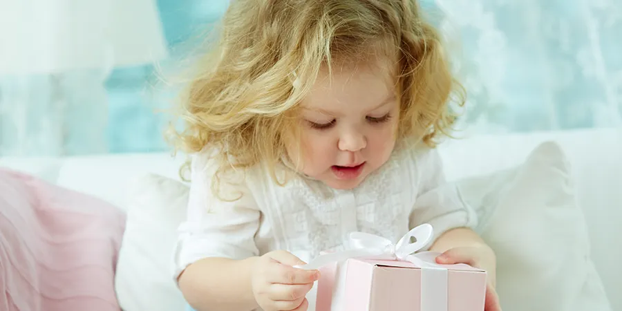Preslatka devojčica, duge, plave kose, uzbuđeno otvara svoj poklon.