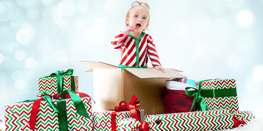 Slatka devojčica, plave kose, prerušena u Deda Mrazovog pomoćnika stoji u kutiji među gomilim upakovanih poklona.