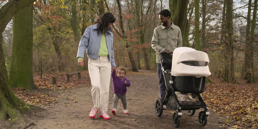 Mladi bračni par šeta sa malim detetom u parku po jesenjem danu. Mama drži ćerku za ruku, dok tata gura luksuzna kolica za decu.