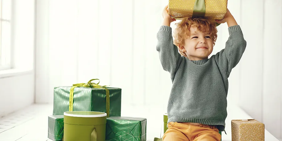 Radosni dečačič sedi na belom drvenom podu, pored gomile lepo upakovanih poklona. Jedan od poklona drži u rukama iznad glave.