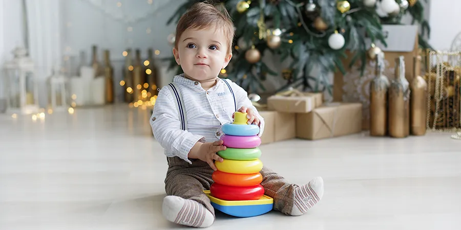 Mali dečak sedi na podu sobe. U pozadini se vidi novogodišnja jelka. Među nogama drži šarenu edukativnu igračku.