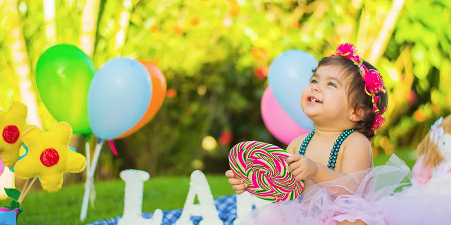 mala devojčica, okružena šarenim balonima, slavi rođendan u parku. U ruci drži šarenu igračku.