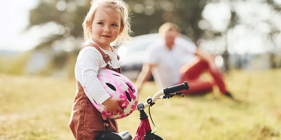 Krupni kadar dvogodišnje devojčice dok pridržava biciklu, sa kacigom u ruci. Ponosni otac je gleda dok sedi na travi u pozadini.