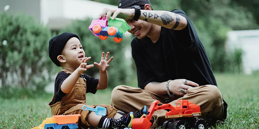 Urbano obučeni otac i sin, sede u travi i igraju se plastičnim autićima.