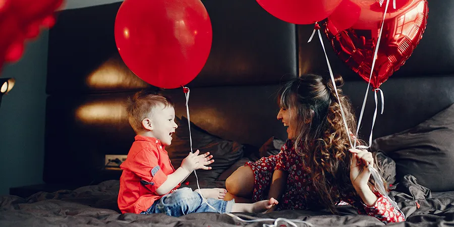 Mali dečak i njegova majka leže u krevetu prekrivenom tamnom posteljinom, dok drže crvene balone.