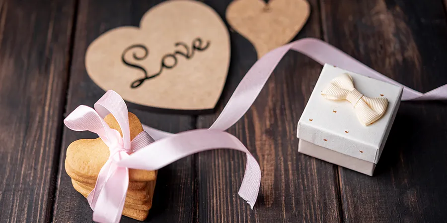 Kolačići u obliku srca, vezani ukrasnom roze mašnom, i lepo upakovani mali poklon se nalaze na drvenom stolu. U sredini, malo iznad, se nalazi drveno srce sa natpisom "love".