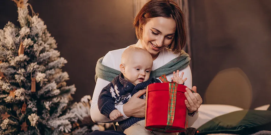 Mama i bebe slave Božićne praznike. Beba sedi mami u krilu, dok mama drži prelepo upakovani poklon crvene boje.