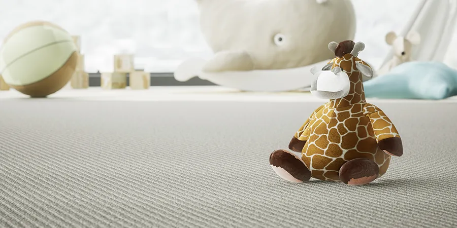 Mala plišana žirafa na sivom tepihu. U pozadini se nazire veliki plišani kit.