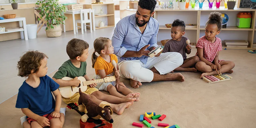 Čovek i petoro male dece, sede na podu u vrtiću dok sviraju muzičke instrumente.