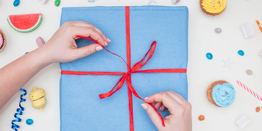 Kadar odozgo. Poklon upakovan u plavi papir sa crvenom trakom. Ženske ruke završavaju vezivanje trakice. Oko poklona se nalaze šarene figurice u obliku kolača i voća.