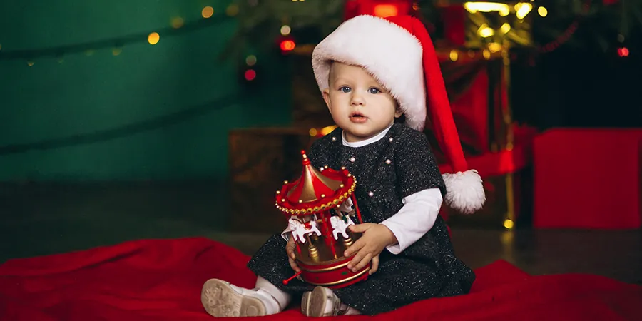 Beba sa novogodišnjom kapicom sedi ispred jelke i drži igračku.