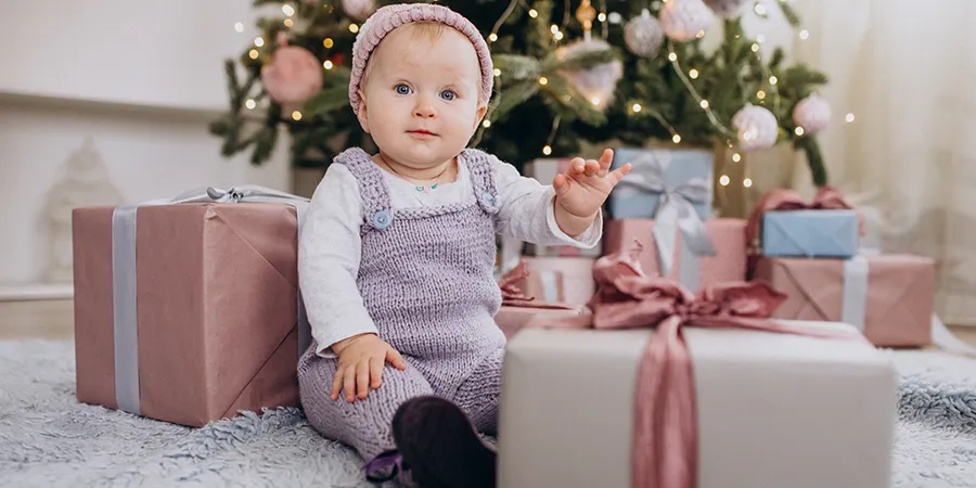 Beba pozira sa poklonima, dok sedi ispred novogodišnje jelke.