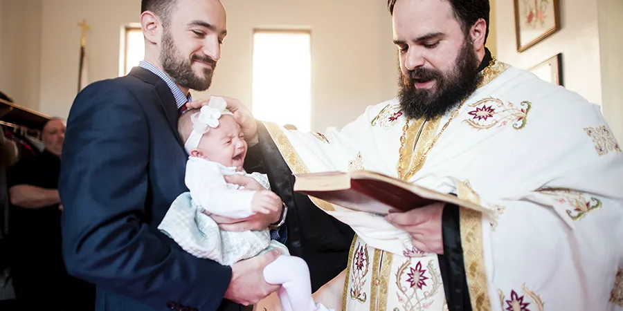 Kum drži malu devojčicu u crkvi, prilikom obreda krštenja. Pop je pored njih, jedna ruka mu je na glavi bebe, dok iz druge čita crkvenu knjigu.