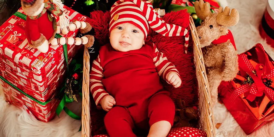 Krupni kadar male bebe u kostimu Deda mrazovog pomoćnika dok leži u drvenoj korpi ispod novogodišnje jelke.