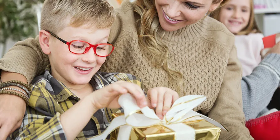 Plavokosi dečak, sa crbenim naočarima za vid, otvara poklon pored svoje mame.