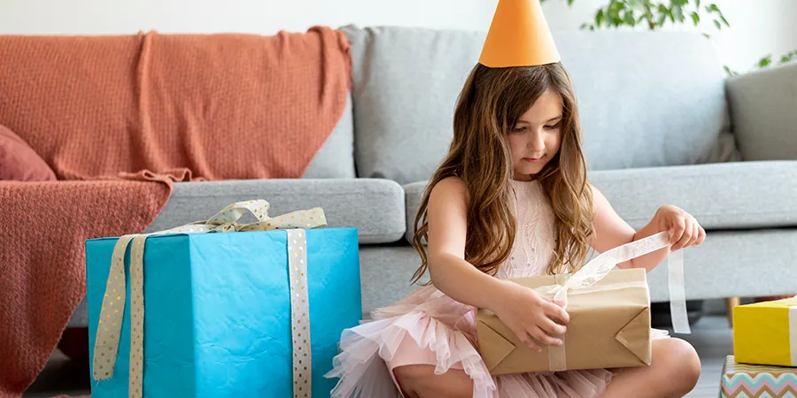 Devojčica sa narandžastom rođendanskom kapicom, sedi na podu i otvara poklone.