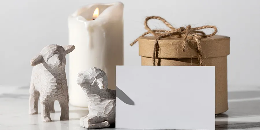 Poklon u retro, kartonskoj kutiji vezan kanapom. Pred poklona je bela velika sveća. Ispred se nalaze prazna čestitka i bele figurice jagnjadi.