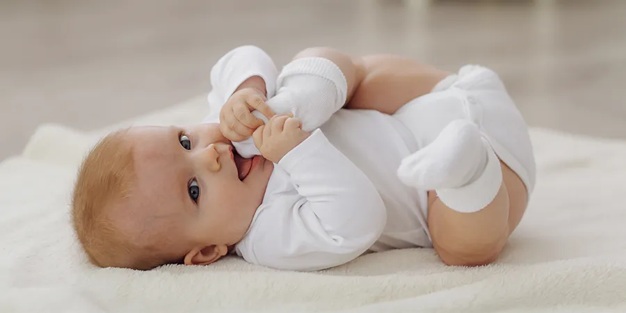 Novorođenče, u beloj odeći, slikano sa strane. Beba stavlja svoja stopala u usta.