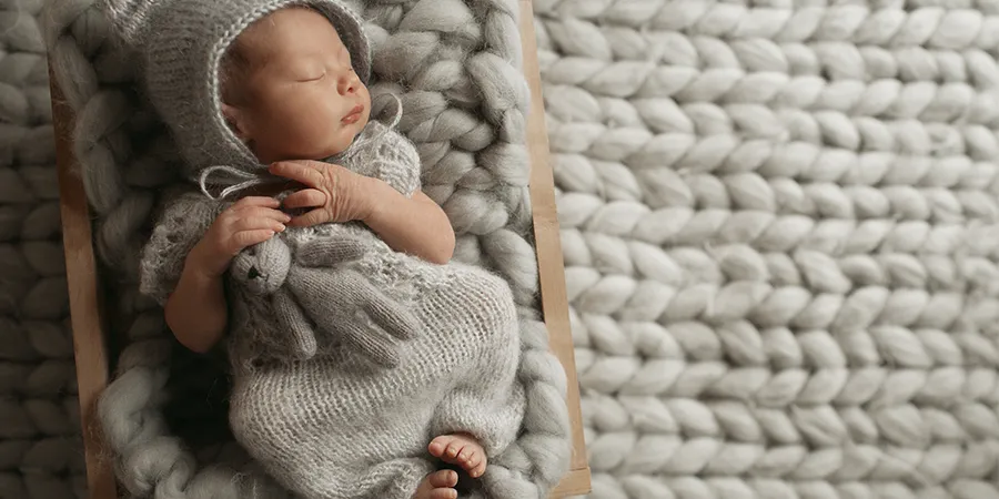 Novorođenče, u sivoj vunenoj odeći sa kapuljačom, na sivom vunenom ćebetu, u drvenoj kolevci. Slika odozgo.