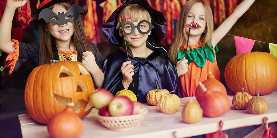Troje dece, sa maskama za Noć veštica, poziraju iza drvenog stola punog bundeva.
