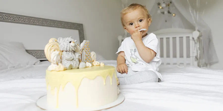 Plavokosi dečačić sedi na bračnom krevetu i gledau kameru dok jede svoju rođendansku tortu. ispred njega he žuta torta sa medom i svećom u obliku broja jedan.