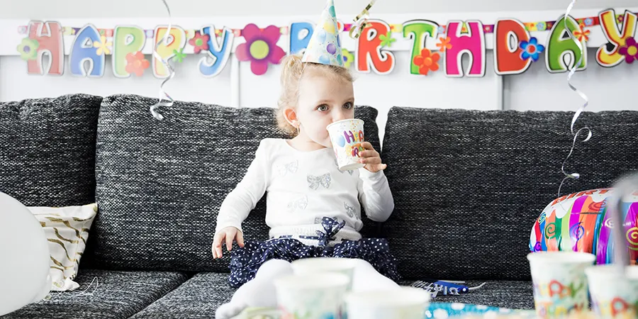 Mala devojčica sa rođendanskom kapoicom na glavi ispija sokić iz čaše dok sedi na sofi. Iza nje na belom zidu je natpis "Happy birthday".