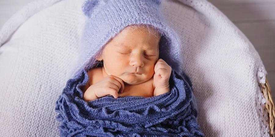 Kadar odozgo novorođene bebe. Beba leži u pletenoj korpi sa belim dušekom i uvijena je u plavo ćebe.