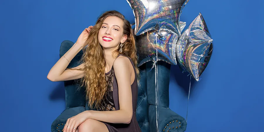 Mlada, nasmejana devoja u sivoj haljini sedi u plavoj fotelji, za koju su vezani baloni. Pozadina je plave boje.