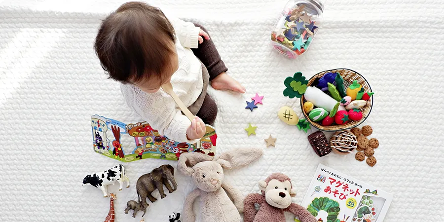 Beba sa drvenom kašikom u ustima, sedi na belom frotiru, dok je okružena raznim igračkama. Slika odozgo.