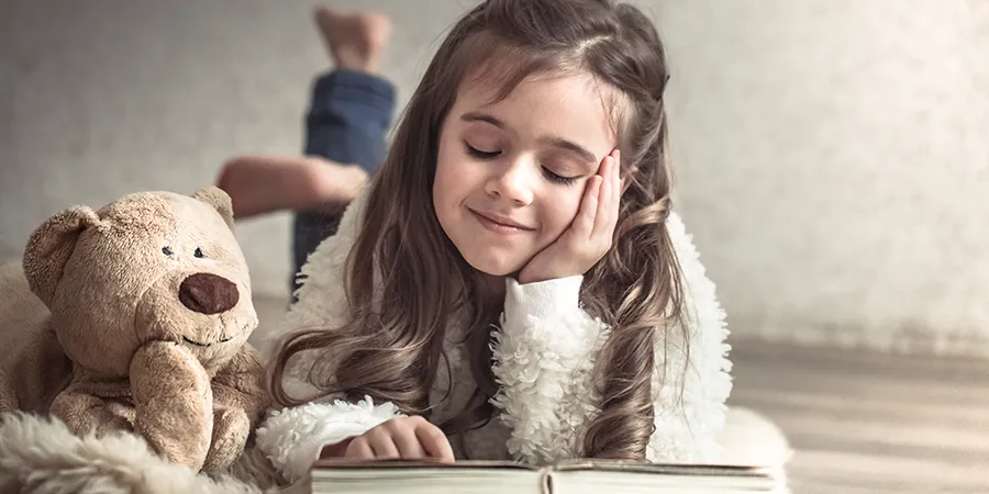 Devojčica leži na drvenom podu dok uživa čitajući svoju omiljenu knjigu. Pored nje se nalazi plišani meda.
