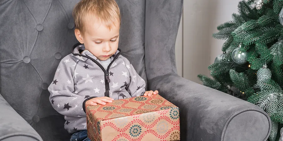 Dečak sedi u sivoj fotelji i drži lepo upakovani poklon u krilu.