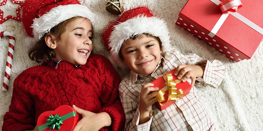 Devojčica i dečak sa novodišnjim kapicama, leže na podu okruženi poklonima.