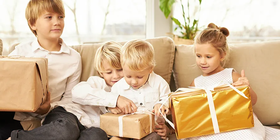 Četvoro dece, plave kose, obučeno u bele košulje, sede na sofi i otvaraju svoje poklone.