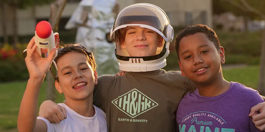 Tri drugara, zagrljeni, poziraju napolju za fotografiju. Dečak u sredini nosi astronautsku kacigu.