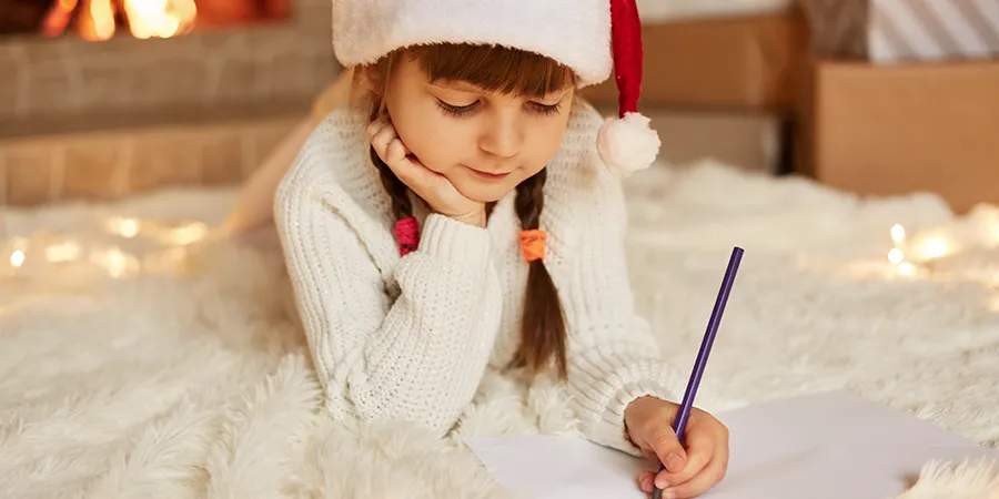 Devojčica, sa novogodišnjom kapom na glavi, piše pismo Deda Mrazu dok leži na podu.
