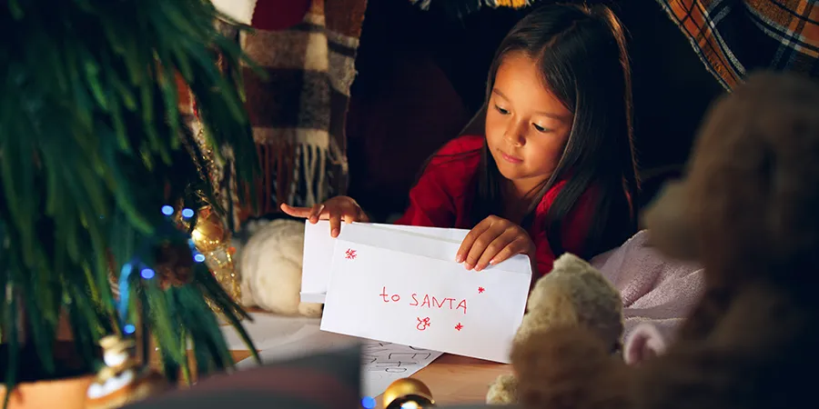 Devojčica koja leži pod novogodišnjom jelkom, pakuje kovertu adresiranu na Deda Mraza.