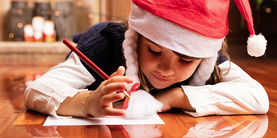 Devojčica, sa novogodišnjom kapom na glavi, piše pismo Deda Mrazu.