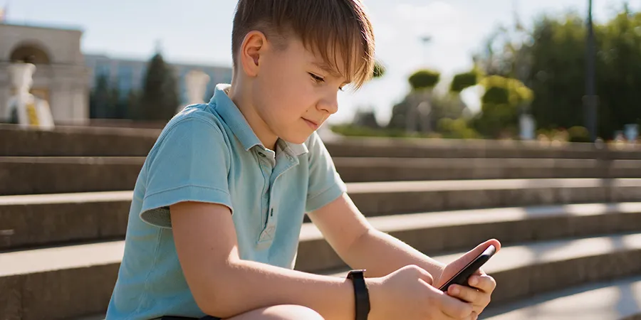 Krupni kadar dečaka koji sedi u parku sa telefonom i pametnim satom u rukama.