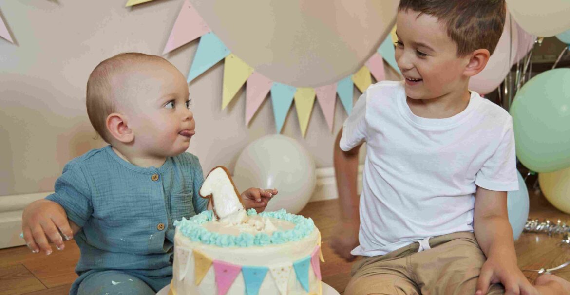 Dvoje male dece sede pored rođendanske torte i smeju se tokom proslave