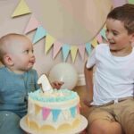 Dvoje male dece sede pored rođendanske torte i smeju se tokom proslave