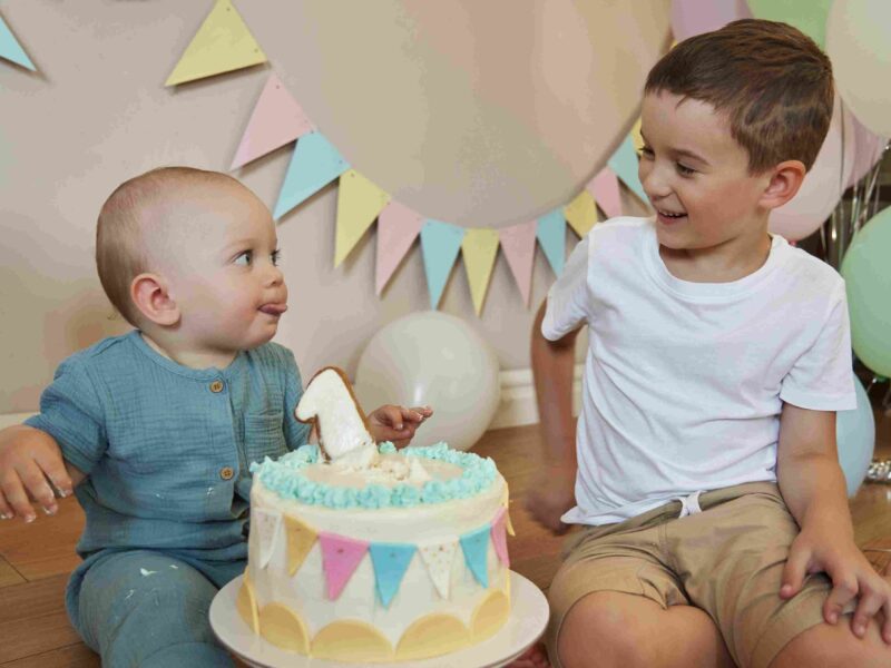 Dvoje male dece sede pored rođendanske torte i smeju se tokom proslave